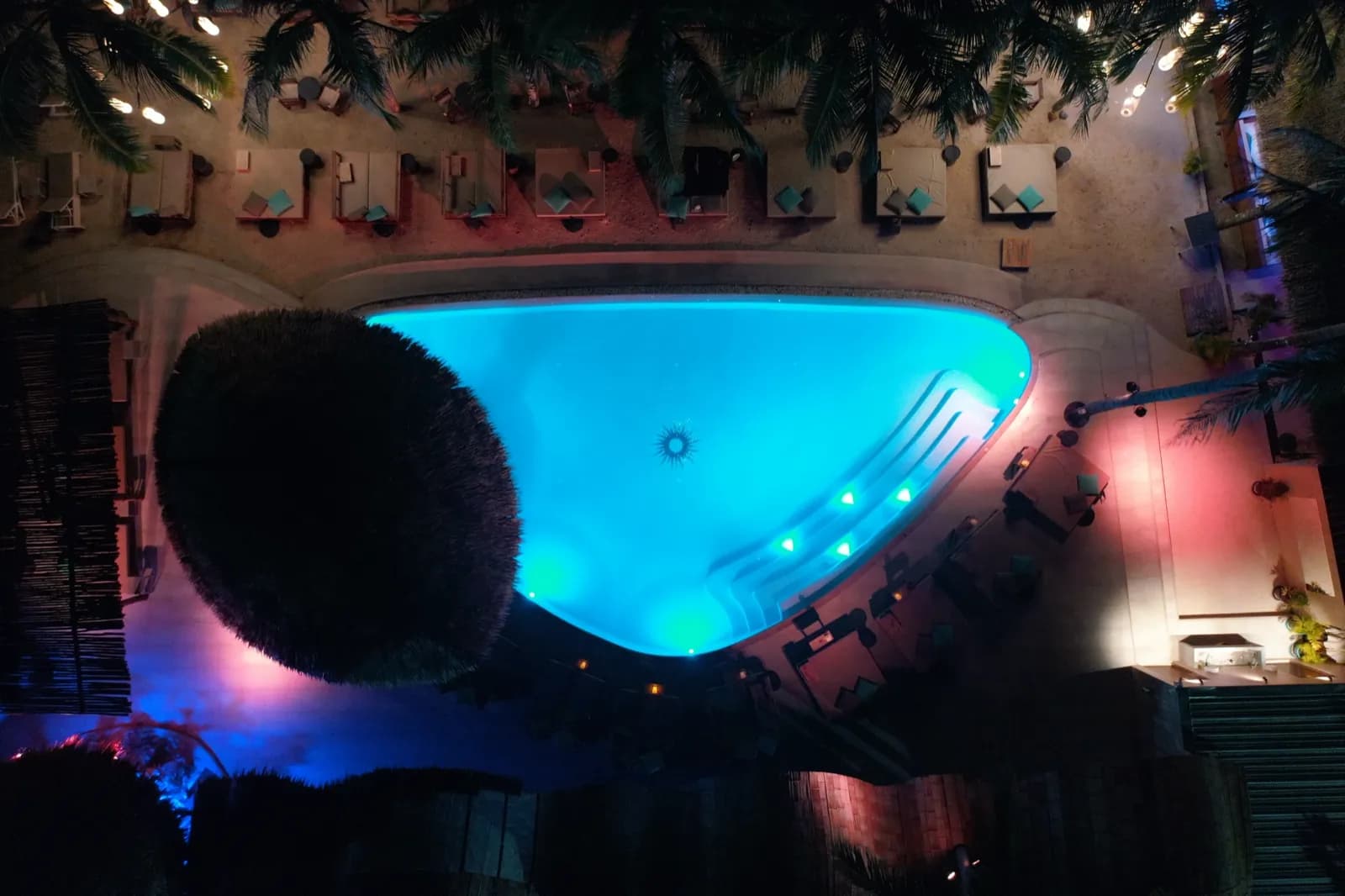 Beach club at night with the pool glowing cyan among pink-lit cabanas