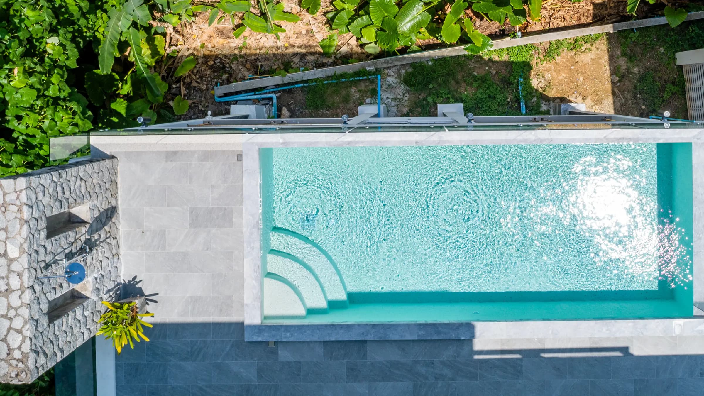 Top-down drone view of a turquoise pool with curved steps beside a stacked-stone shower wall