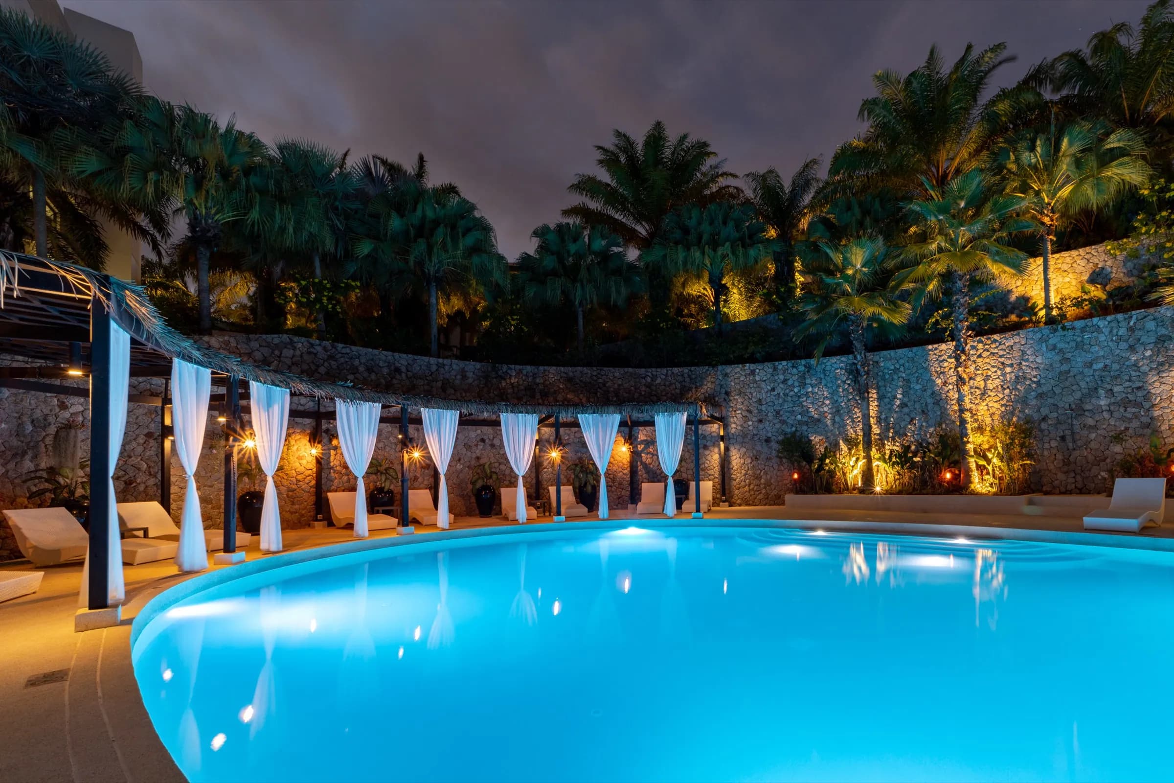 Illuminated resort pool at night before curtained cabanas and a stone wall