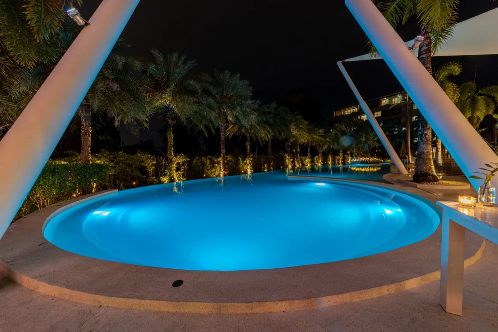 Round plunge pool lit cyan at night between white sail-shade masts
