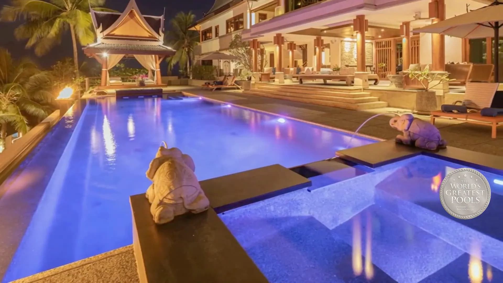 Thai villa and infinity pool at blue hour with glowing fire bowls and violet pool lights