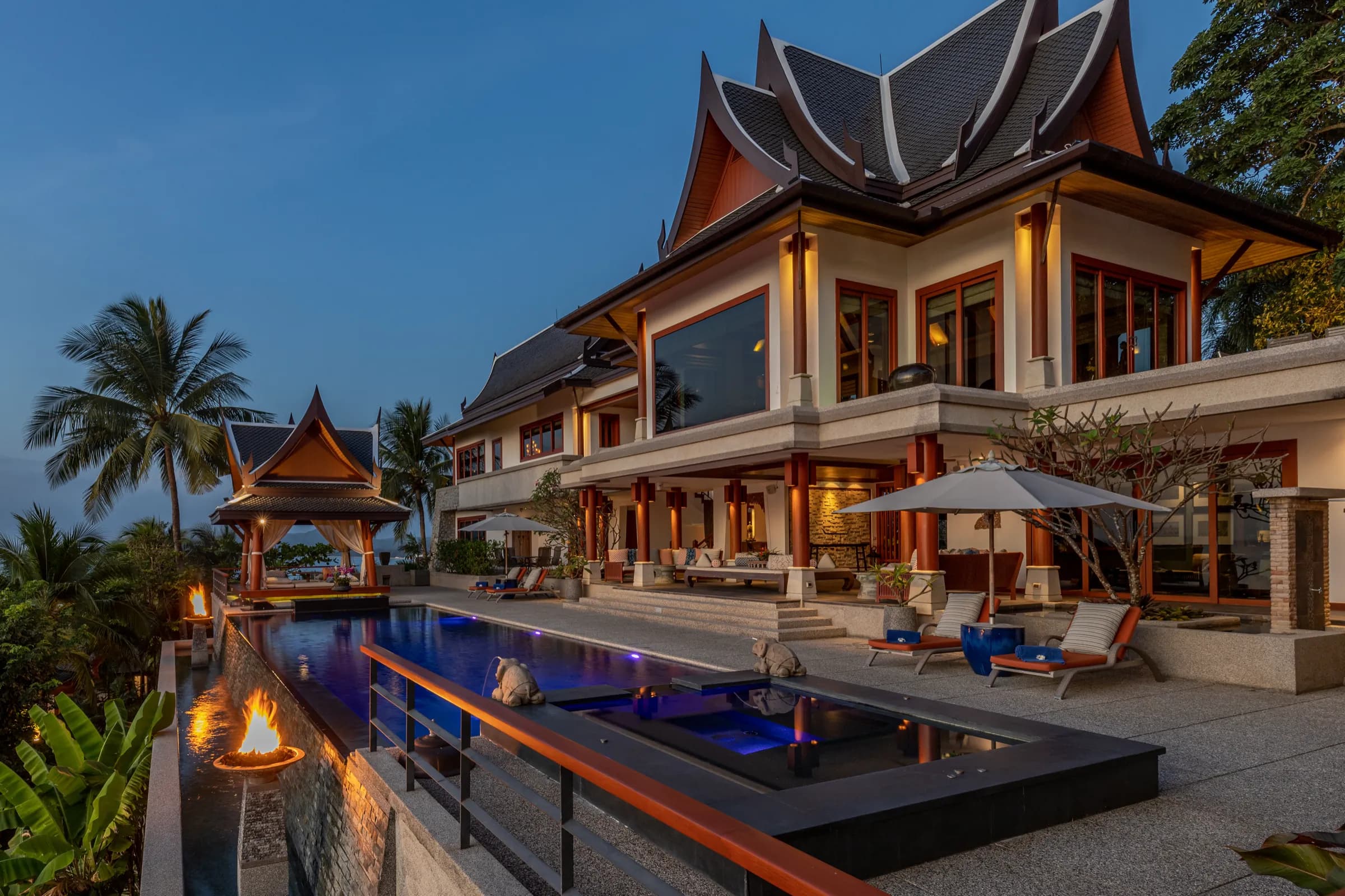 Thai-style villa and long infinity pool at blue hour, with fire bowls along the edge