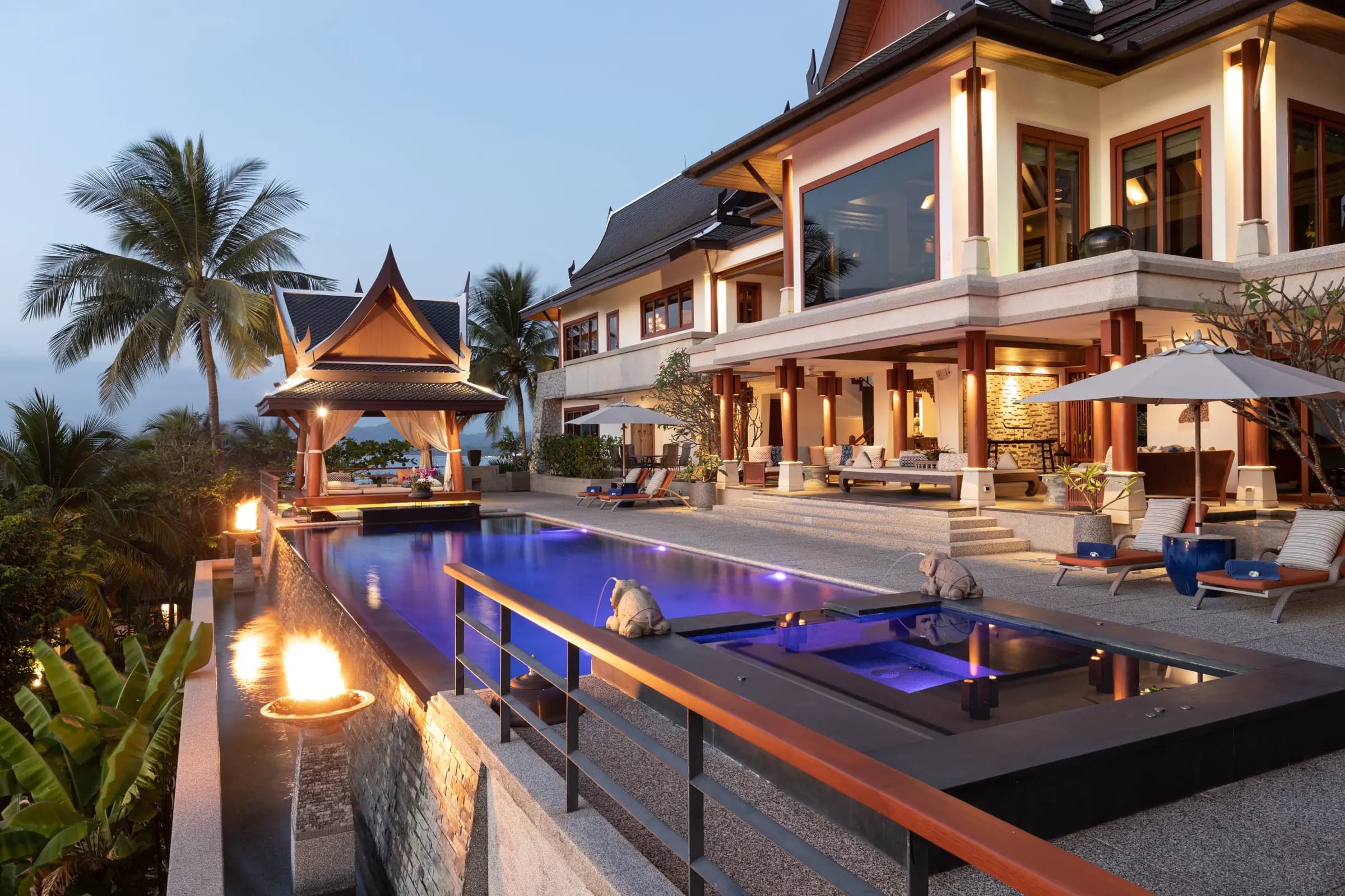 Thai villa and infinity pool at blue hour with glowing fire bowls and violet pool lights